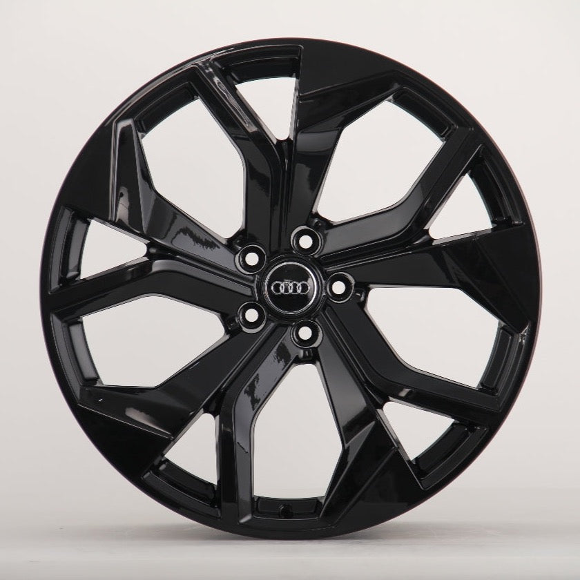 A glossy black alloy wheel with a multi-spoke design. The Audi logo is centered on the hub. It's set against a plain, light grey background.