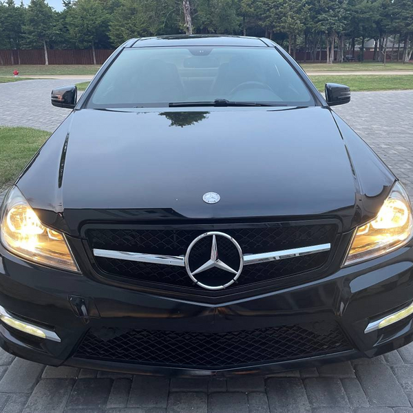 A black sedan with headlights on, parked on a paved driveway. It features a prominent emblem on the grille, surrounded by a residential area with trees and grass.