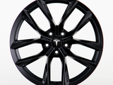 A black wheel rim featuring a sleek, multi-spoke design with the Tesla logo visible at the center, placed against a plain white background.