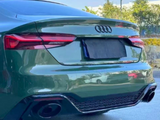 A green Audi car is parked, displaying its rear with dual exhaust pipes. Surrounding it is a paved area with trees and clear blue sky in the background.