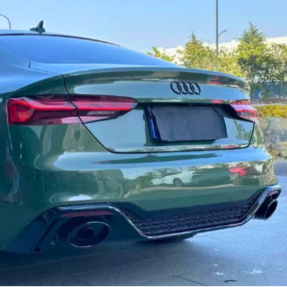 A green Audi car is parked, displaying its rear with dual exhaust pipes. Surrounding it is a paved area with trees and clear blue sky in the background.
