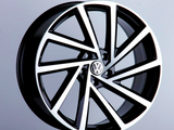 A stylish black and silver alloy wheel, featuring the Volkswagen logo, is displayed upright on a dark stand against a plain gray background.