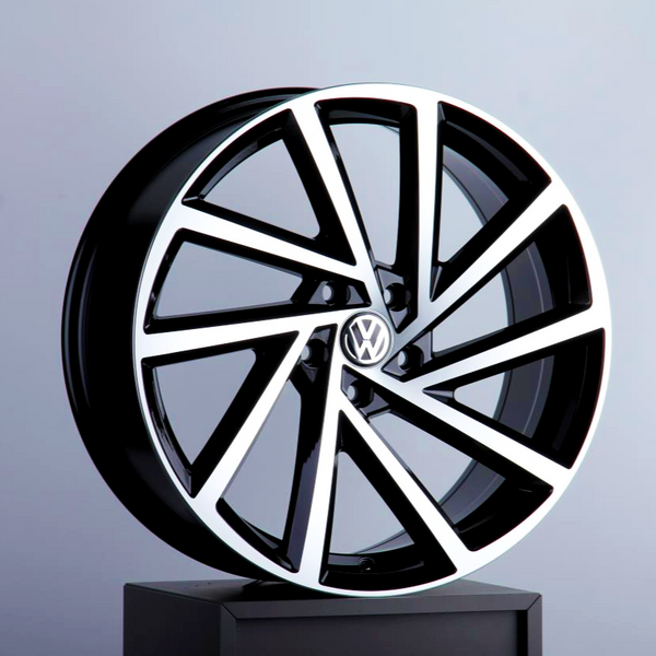 A stylish black and silver alloy wheel, featuring the Volkswagen logo, is displayed upright on a dark stand against a plain gray background.