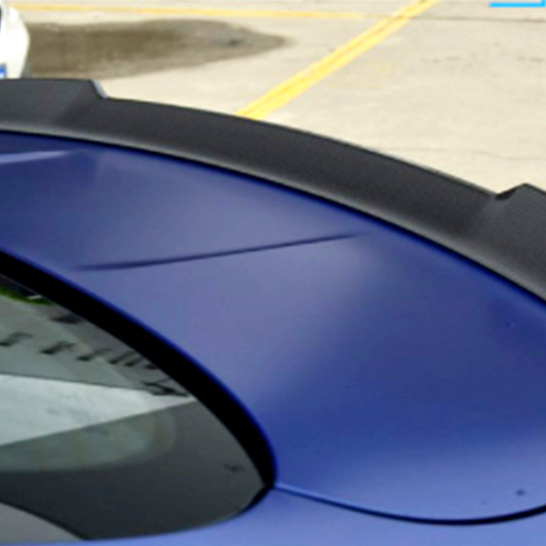 A black carbon fiber spoiler sits on the rear of a blue car, with a white car parked nearby in an outdoor concrete area marked by yellow lines.