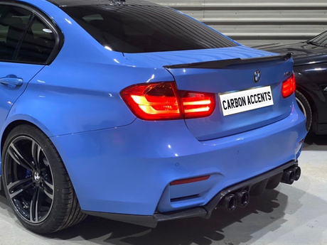A blue car with illuminated taillights is parked in an indoor garage. The license plate reads, "CARBON ACCENTS." The car has dual exhausts and a carbon-fiber rear spoiler.