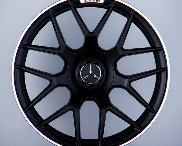 A black multi-spoke car wheel rim with a Mercedes-Benz logo at the center, labeled "AMG," is displayed against a neutral background, resting atop a pedestal.
