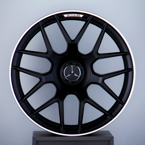 A black multi-spoke car wheel rim with a Mercedes-Benz logo at the center, labeled "AMG," is displayed against a neutral background, resting atop a pedestal.