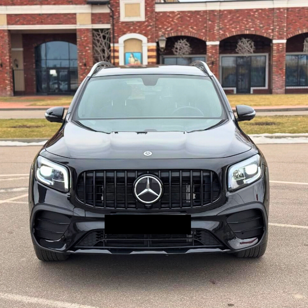 A black Mercedes SUV is parked, facing forward, in an outdoor lot with a brick building backdrop. The car's lights are on, and it features distinctive grille and emblem design.