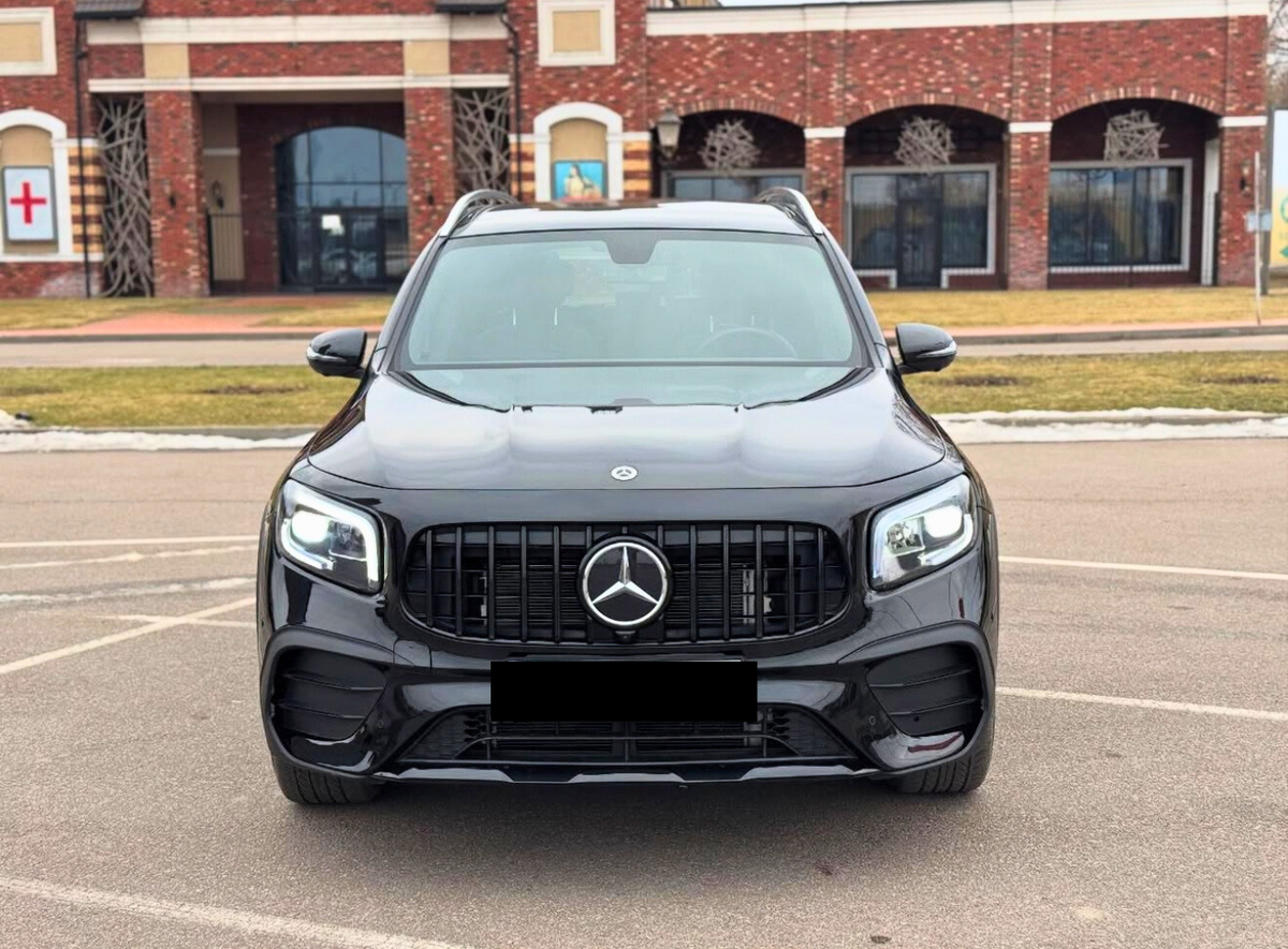 A black Mercedes SUV is parked, facing forward, in an outdoor lot with a brick building backdrop. The car's lights are on, and it features distinctive grille and emblem design.