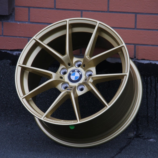 A gold BMW wheel rim rests against a brick-patterned wall on asphalt. The center features the BMW logo with spokes radiating outward.