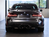 A sleek, dark-colored BMW car is stationary, viewed from behind. It's indoors in a brightly lit space with large windows, showing its signature quad exhaust pipes and prominent rear spoiler.