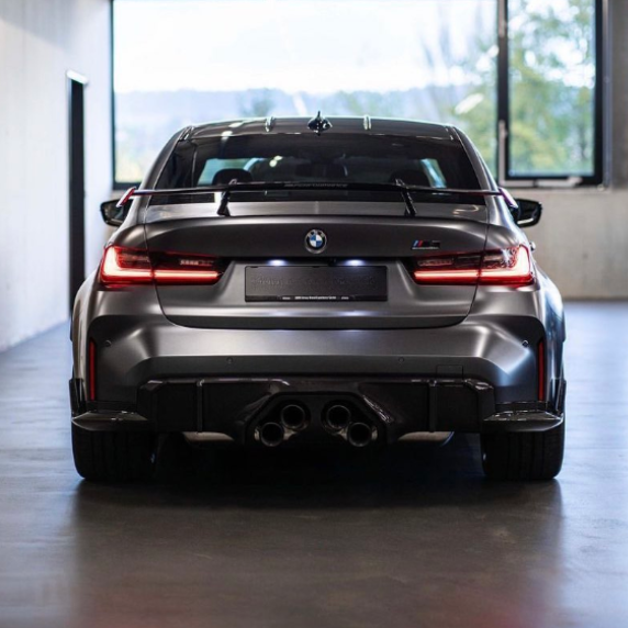 A sleek, dark-colored BMW car is stationary, viewed from behind. It's indoors in a brightly lit space with large windows, showing its signature quad exhaust pipes and prominent rear spoiler.
