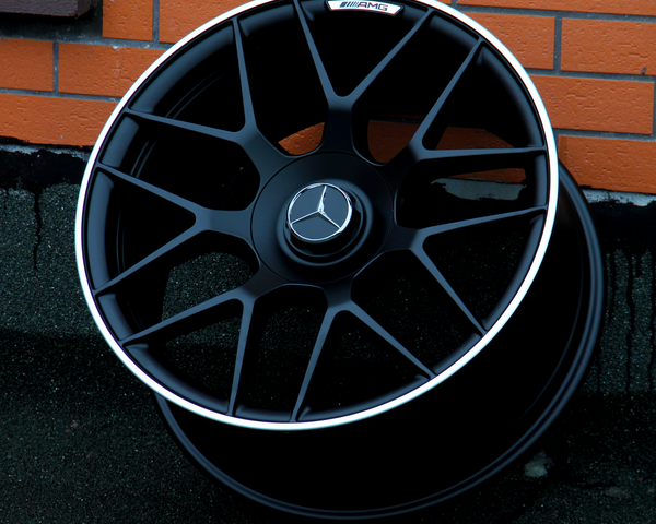 A black AMG car wheel rests against an orange brick wall. The wheel features a Mercedes logo at its center.