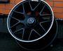A black AMG car wheel rests against an orange brick wall. The wheel features a Mercedes logo at its center.