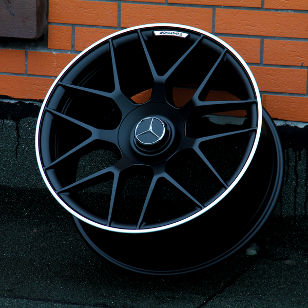 A black AMG car wheel rests against an orange brick wall. The wheel features a Mercedes logo at its center.
