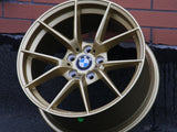 A gold BMW alloy wheel rests against a red brick wall, showcasing its multi-spoke design. The BMW logo is centered, surrounded by bolt holes. The surface is smooth and polished.
