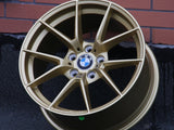 A gold BMW alloy wheel rests against a brick wall on an asphalt surface. The BMW logo is centered on the wheel's hub.