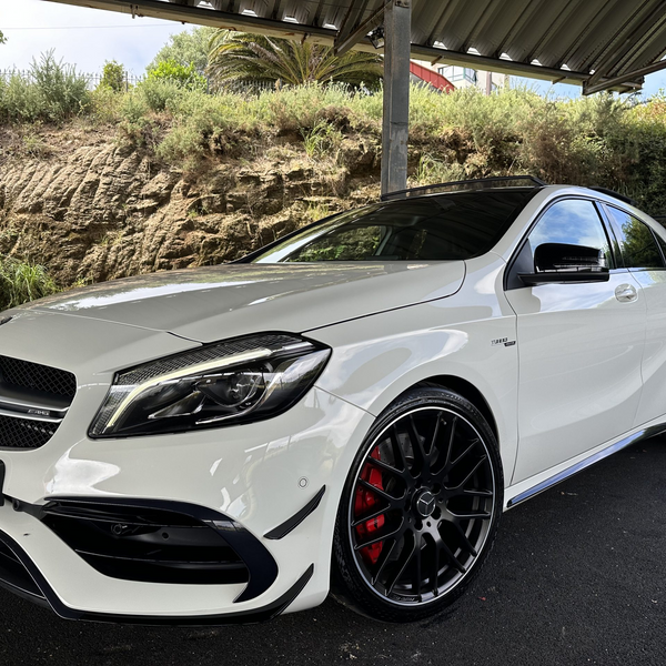 A white Mercedes-Benz sedan is parked under a metal-roofed structure, featuring sleek black wheels and set against a backdrop of rocks and dense shrubs.