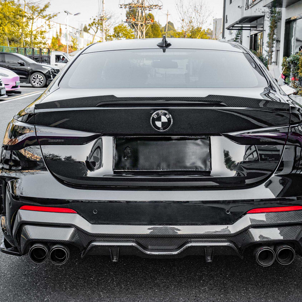 A black BMW car parked on a street, viewed from behind, surrounded by trees and other parked vehicles. The environment is urban, with buildings visible on the right.