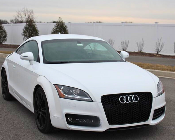 A white Audi car, parked on a paved road, displays a prominent logo on the front grille. The background features a white fence and sparse trees against a cloudy sky.