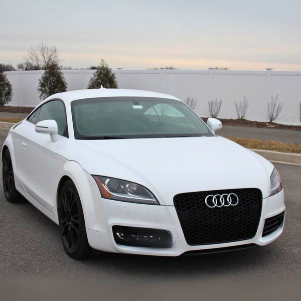 A white Audi car, parked on a paved road, displays a prominent logo on the front grille. The background features a white fence and sparse trees against a cloudy sky.