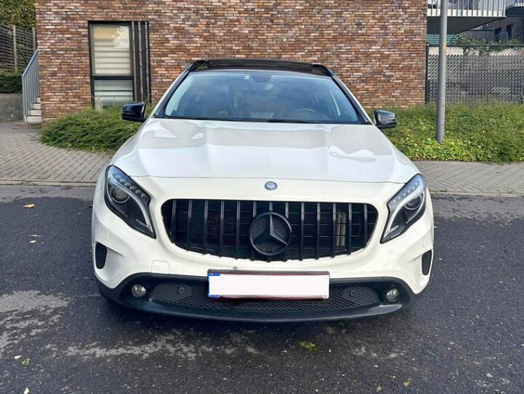 A white Mercedes-Benz SUV is parked on a paved street. The background shows a brick building and greenery, with a sidewalk lining the scene.