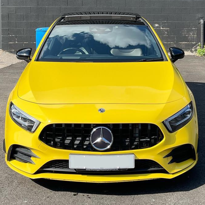 A bright yellow Mercedes-Benz parked on an asphalt surface, reflecting clouds on its windshield, is positioned against a black brick wall. A blue bin is partially visible beside it.