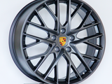 A dark metallic alloy wheel with multiple spokes and a central emblem featuring the Porsche logo, displayed on a platform against a plain white background.