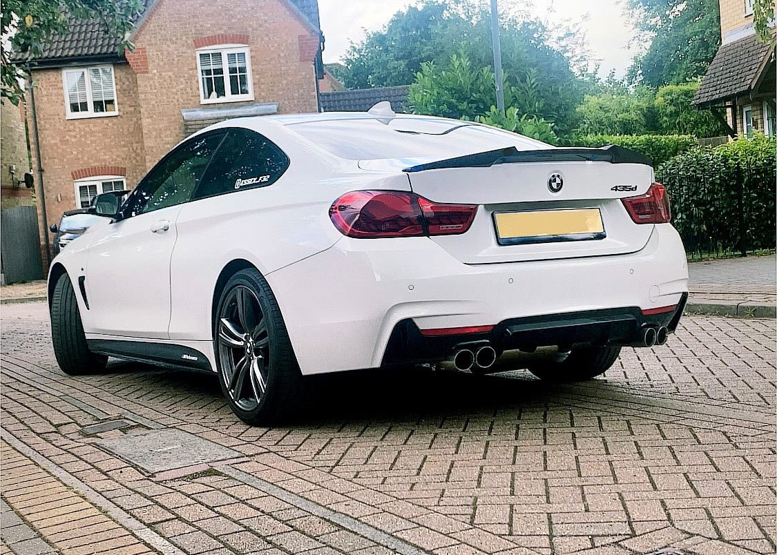 A white BMW 435d is parked on a brick driveway in a residential area, featuring distinctive dual exhausts and surrounded by houses and greenery.