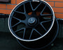 A black Mercedes-Benz AMG wheel rests against a brick wall on a dark, textured ground. The wheel features a silver trim and the Mercedes-Benz logo at the center.
