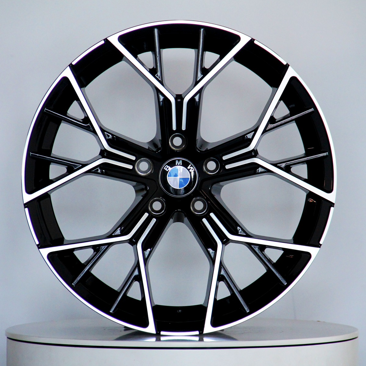 A black and silver BMW alloy wheel, featuring a multi-spoke design, is displayed upright on a light gray circular platform. The BMW logo is centered.