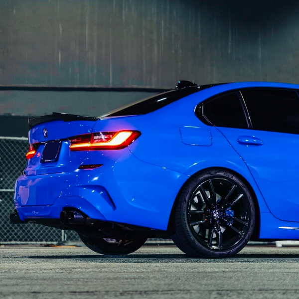 A blue sedan with illuminated taillights is parked on a concrete surface, surrounded by a chain-link fence and under an industrial-style structure.