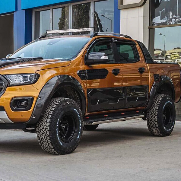A rugged orange pickup truck with large off-road tires is parked beside a modern building, featuring a roof light bar and black accents.