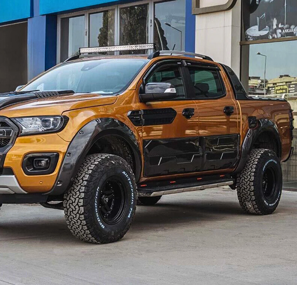 A rugged orange pickup truck with large off-road tires is parked beside a modern building, featuring a roof light bar and black accents.