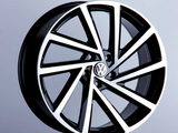 A black and silver alloy car wheel, featuring a Volkswagen logo at the center, is displayed upright on a pedestal against a plain, light gray background.