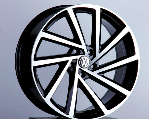 A black and silver alloy car wheel, featuring a Volkswagen logo at the center, is displayed upright on a pedestal against a plain, light gray background.