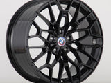 A sleek black alloy car wheel with a geometric spoke design sits against a plain white background. The wheel features a BMW logo at its center.