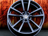 A black, multi-spoke Volkswagen wheel rim rests against an orange, textured background on a gray surface. The Volkswagen logo is prominently displayed in the center.