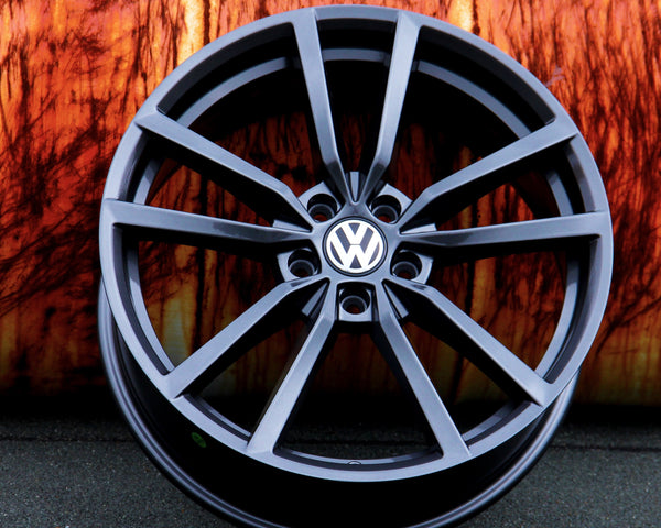 A black, multi-spoke Volkswagen wheel rim rests against an orange, textured background on a gray surface. The Volkswagen logo is prominently displayed in the center.