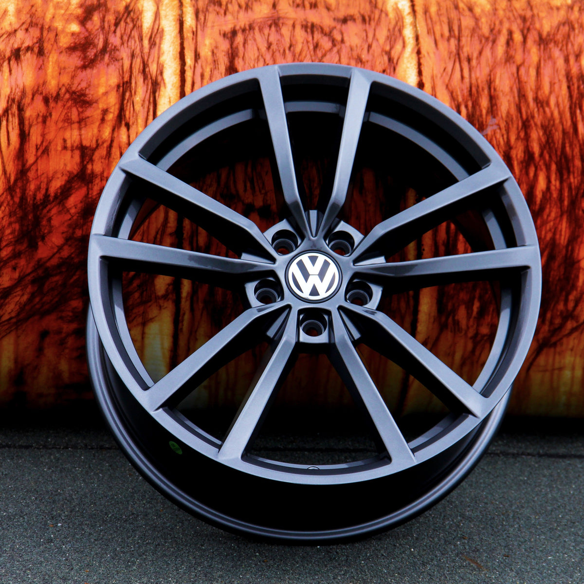 A black, multi-spoke Volkswagen wheel rim rests against an orange, textured background on a gray surface. The Volkswagen logo is prominently displayed in the center.