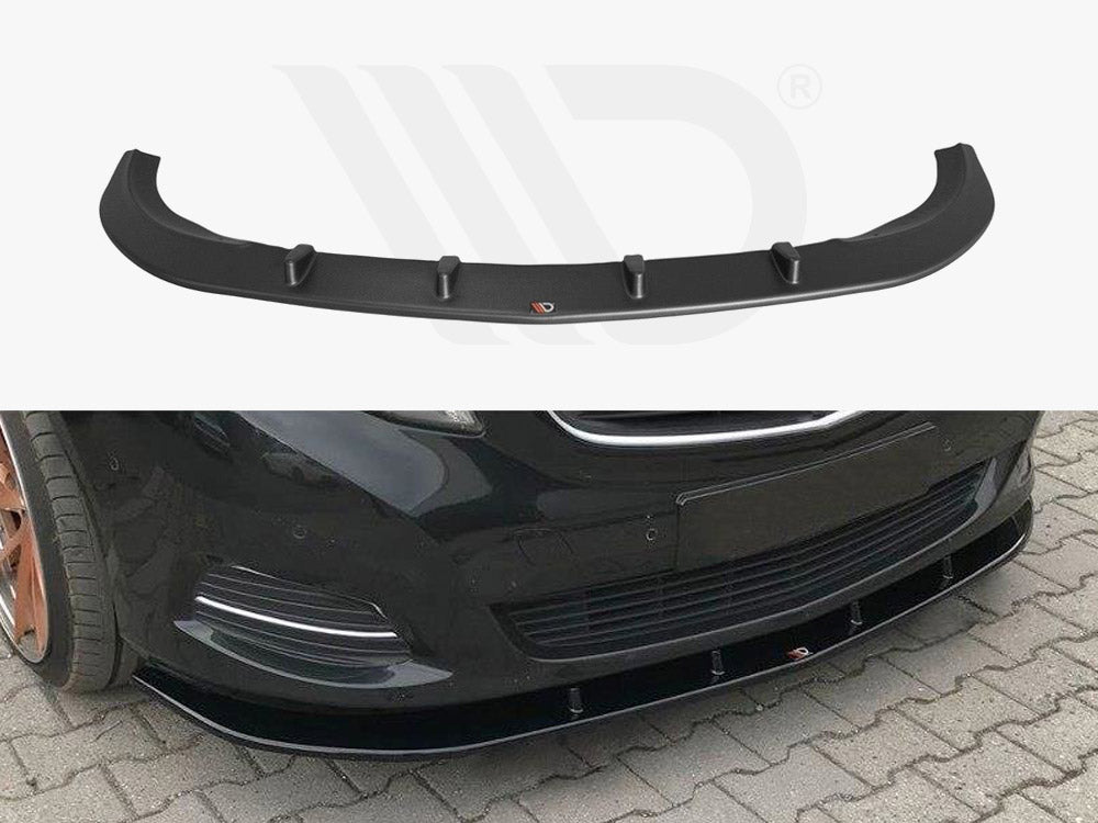 A black car front spoiler with aerodynamic fins is displayed. It is positioned on the front bumper of a black car parked on a cobblestone surface.