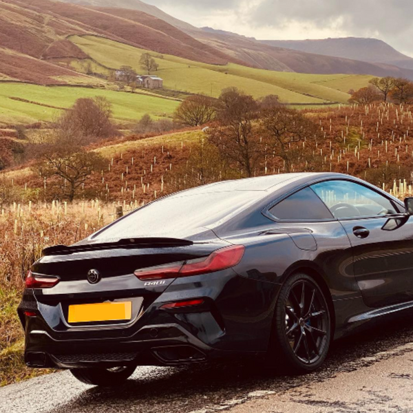 A black car is parked on a rural road, surrounded by rolling green hills and trees under a cloudy sky. Car badge reads "840i."
