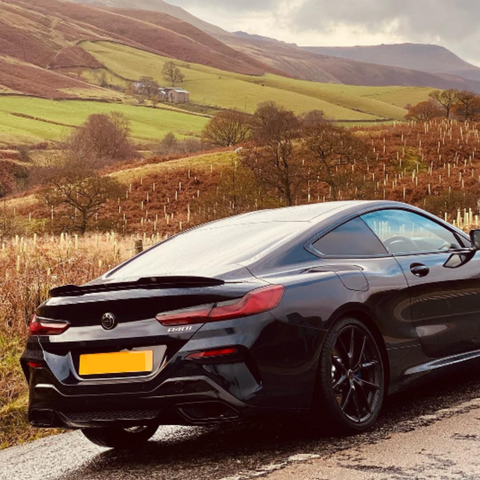 A black car is parked on a rural road, surrounded by rolling green hills and trees under a cloudy sky. Car badge reads "840i."