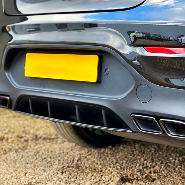 The rear end of a dark car is parked on gravel, featuring a bright yellow license plate, dual exhausts, and red reflectors. Nearby, other vehicles are visible in the reflection.