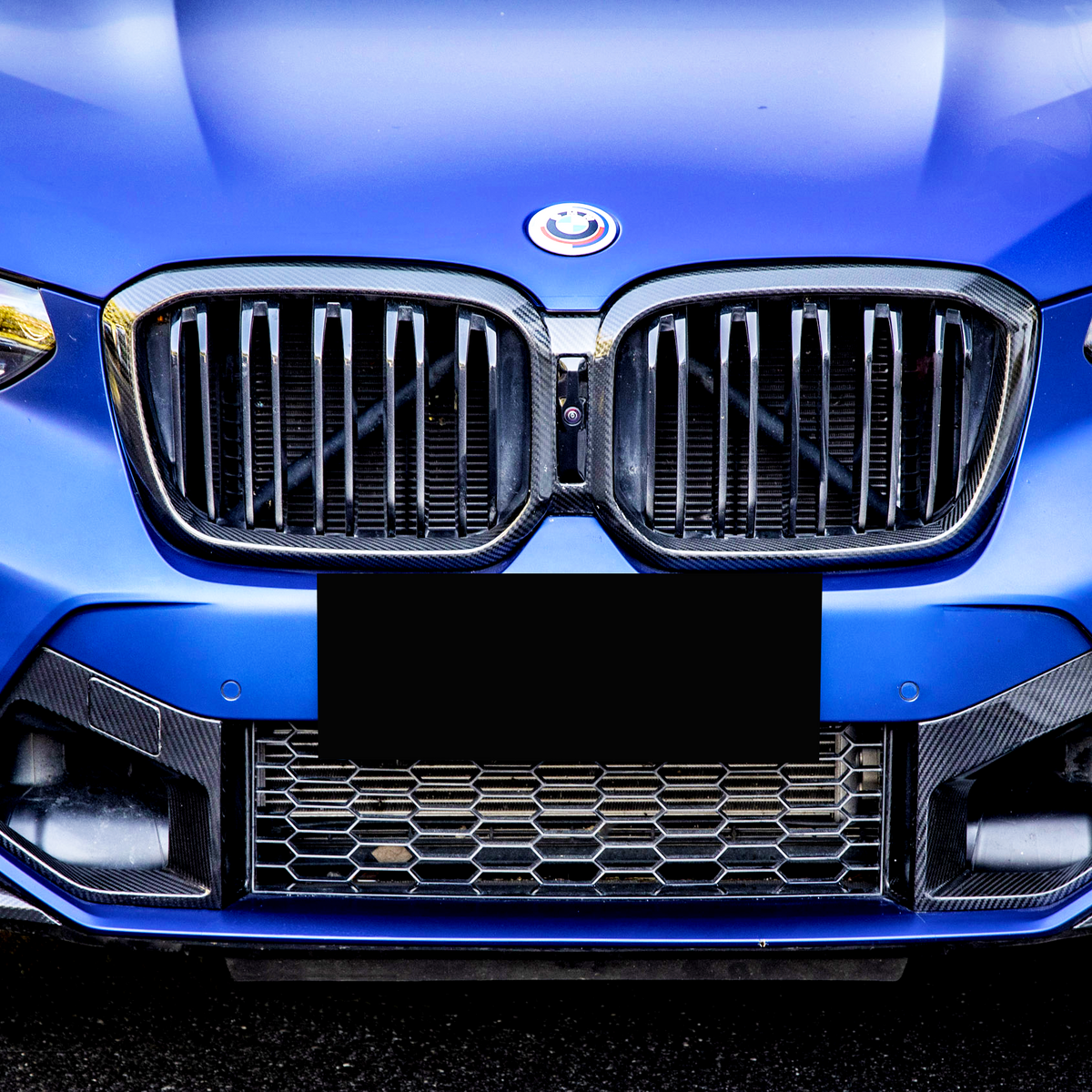 A blue car's front features a modified grille with a smiling logo, surrounded by bright headlights. The vehicle is parked against a blurred urban background.