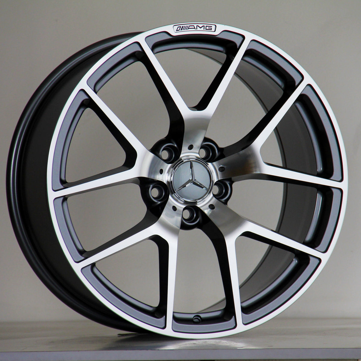 A Mercedes-Benz alloy wheel stands upright, featuring a five-spoke design with a polished finish. The AMG logo is inscribed at the top. It's set against a neutral background.