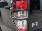 A rear vehicle taillight with red and white sections is mounted on a glossy dark surface, reflecting an urban environment with buildings. The letter 