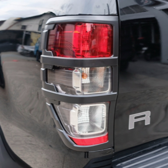 A rear vehicle taillight with red and white sections is mounted on a glossy dark surface, reflecting an urban environment with buildings. The letter "R" appears nearby.