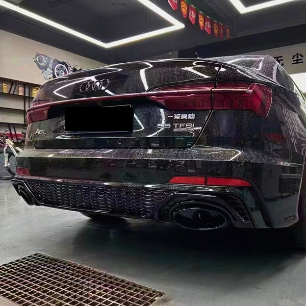 A sleek black Audi A6L with dual exhausts is parked indoors under bright, square ceiling lights. Wall art and a shelf with trophies are visible. Text: "A6L", "55 TFSI".