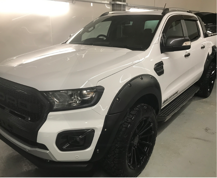 White pickup truck parked indoors, showcasing a robust design with large tires and a black grille. The side has "WILDTRAK" branding. The surrounding environment is a clean garage.